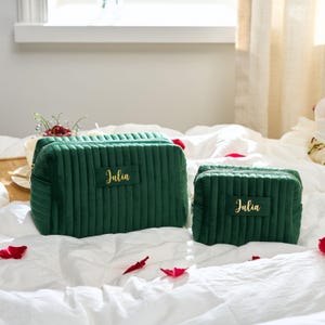 May include: Two emerald green velvet cosmetic bags with vertical ribbed detailing. The larger bag has a rectangular label with the name "Julia" in gold script. The smaller bag also has the name "Julia" in gold script.
