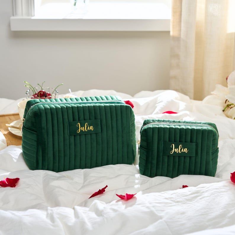 May include: Two emerald green velvet cosmetic bags with vertical ribbed detailing. The larger bag has a rectangular label with the name "Julia" in gold script. The smaller bag also has the name "Julia" in gold script.