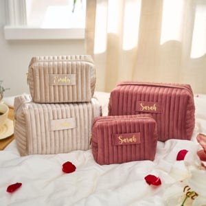 May include: Four personalized cosmetic bags in varying sizes and colors. Two are beige, and two are a dusty rose color. Each bag has a gold zipper and a rectangular patch with a name in gold lettering. The bags are arranged on a white surface.
