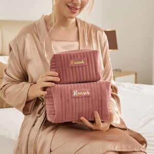 May include: Two dusty rose quilted cosmetic bags with the name "Hannah" in gold lettering. The bags are rectangular with a zipper closure. The bags are different sizes and are stacked on top of each other.