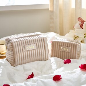 May include: Two beige, corduroy-textured cosmetic bags with gold zippers and the name "Emily" in gold lettering. The bags are on a white surface with red rose petals scattered around them. The larger bag is rectangular, and the smaller bag is square.