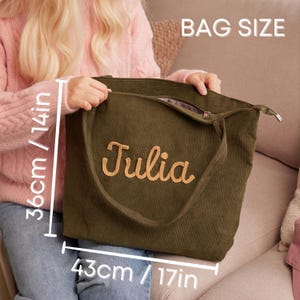 May include: Olive green corduroy tote bag with the name "Julia" embroidered in a light brown script. The bag measures 17 inches wide and 14 inches high. The bag has a zipper closure and a shoulder strap. The text "BAG SIZE" is visible.