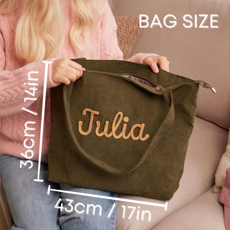 May include: Olive green corduroy tote bag with the name "Julia" embroidered in a light brown script. The bag measures 17 inches wide and 14 inches high. The bag has a zipper closure and a shoulder strap. The text "BAG SIZE" is visible.