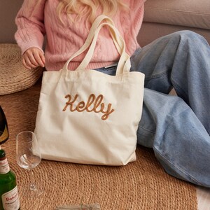 May include: Cream-colored corduroy tote bag with the name "Kelly" embroidered in a brown script. The bag has two handles and is sitting on a woven mat. The background includes a pink sweater and blue jeans.