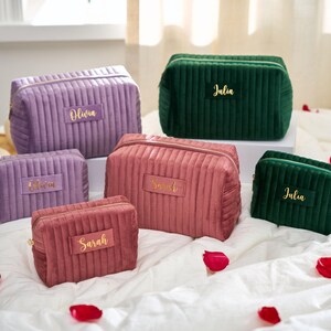 May include: A collection of six personalized cosmetic bags in various colors, including purple, pink, and green. Each bag features a gold nameplate with names like "Olivia" and "Julia". The bags are made of a ribbed, velvet-like material.