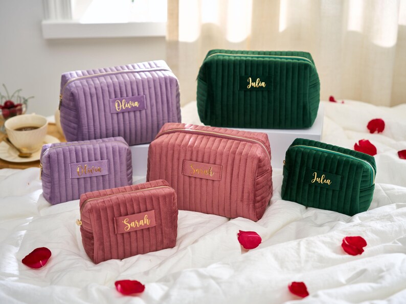 May include: A collection of six personalized cosmetic bags in various colors, including purple, pink, and green. Each bag features a gold nameplate with names like "Olivia" and "Julia". The bags are made of a ribbed, velvet-like material.