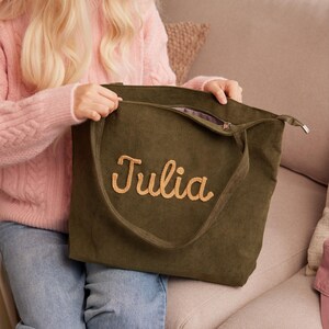 May include: An olive green corduroy tote bag with the name "Julia" embroidered in a light tan color. The bag has a zipper closure and a long strap. The bag is held by a person wearing a pink sweater.