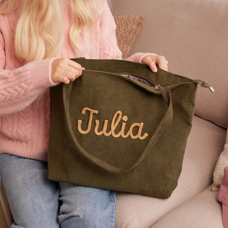 May include: An olive green corduroy tote bag with the name "Julia" embroidered in a light tan color. The bag has a zipper closure and a long strap. The bag is held by a person wearing a pink sweater.