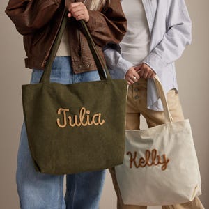 May include: Two corduroy tote bags, one olive green and one cream. The olive green bag has the name "Julia" embroidered in a light brown script. The cream bag has the name "Kelly" embroidered in a light brown script.
