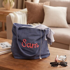 May include: A blue corduroy tote bag with the name "Sam" embroidered in pink. The bag is open, revealing a striped shirt and white earbuds. Sunglasses, perfume, and lip gloss sit on a wooden table.