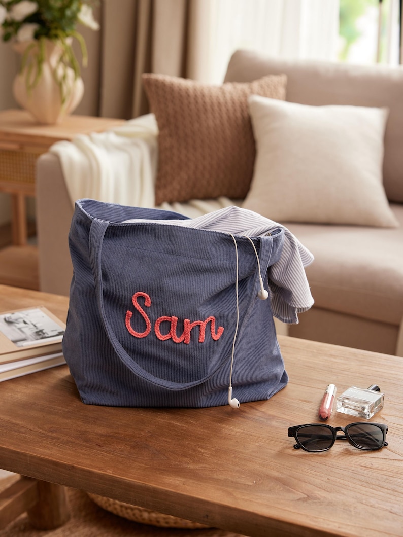 May include: A blue corduroy tote bag with the name "Sam" embroidered in pink. The bag is open, revealing a striped shirt and white earbuds. Sunglasses, perfume, and lip gloss sit on a wooden table.