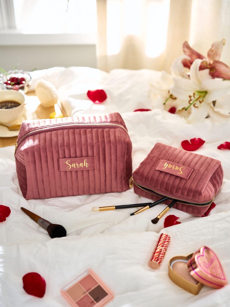 May include: Two dusty rose velvet makeup bags with gold zippers and the name "Sarah" embroidered on them. Makeup brushes, lipstick, and a heart-shaped compact are arranged on a white bedspread. A makeup palette is also visible.