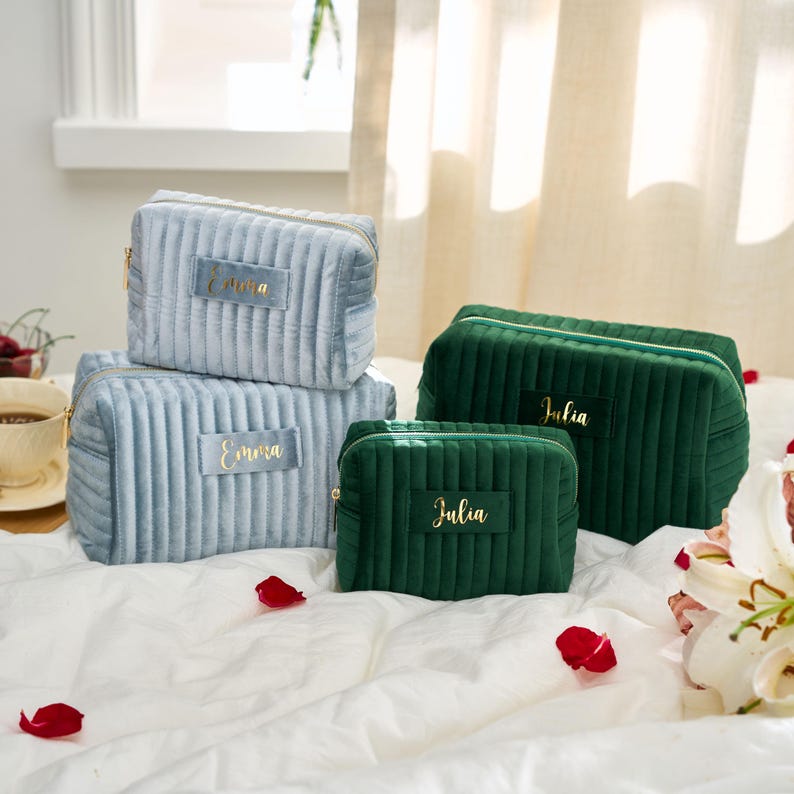 May include: A collection of plush, ribbed cosmetic bags in various sizes and colors. The bags are in shades of blue and green, with gold zippers and personalized name tags. The bags are arranged on a white bedspread.