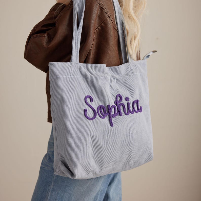 May include: A light blue corduroy tote bag with the name "Sophia" embroidered in purple script. The bag has matching light blue handles and is being carried over the shoulder. The bag is made of a ribbed corduroy fabric.
