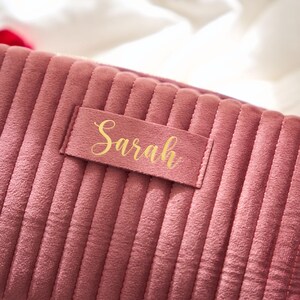 May include: A dusty rose-colored, corduroy makeup bag with vertical ribbed detailing. A rectangular label with the name "Sarah" in gold script is affixed to the front. The bag is designed for cosmetics and personal care items.