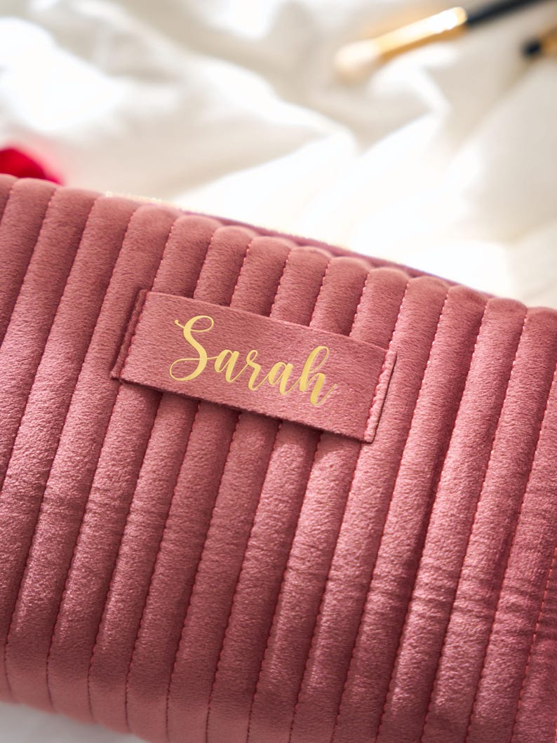 May include: A dusty rose-colored, corduroy makeup bag with vertical ribbed detailing. A rectangular label with the name "Sarah" in gold script is affixed to the front. The bag is designed for cosmetics and personal care items.