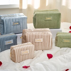 May include: A collection of corduroy cosmetic bags in various sizes and colors, including blue, beige, and green. Each bag features a gold zipper and a rectangular label with a name in gold lettering. The bags are arranged on a white surface with scattered rose petals.