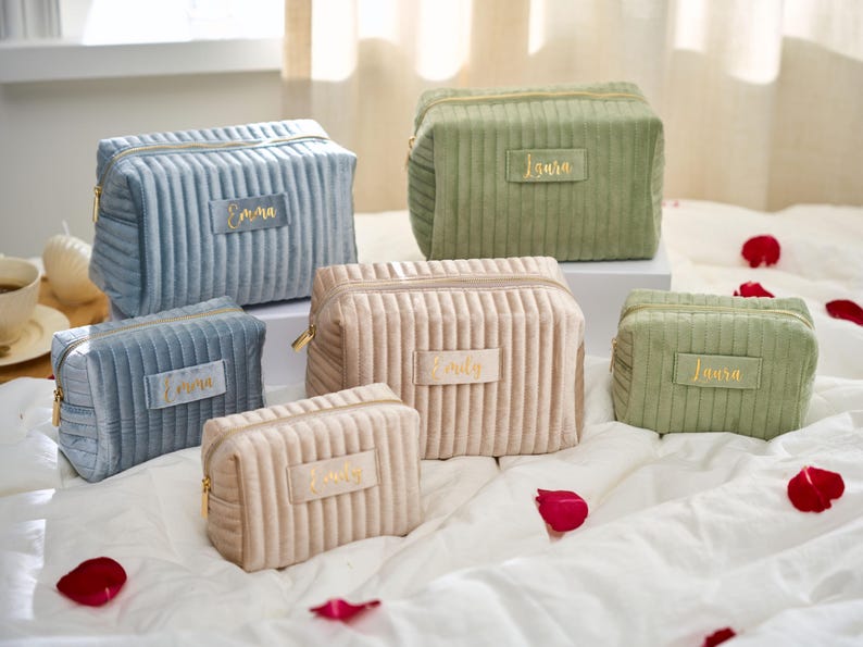 May include: A collection of corduroy cosmetic bags in various sizes and colors, including blue, beige, and green. Each bag features a gold zipper and a rectangular label with a name in gold lettering. The bags are arranged on a white surface with scattered rose petals.