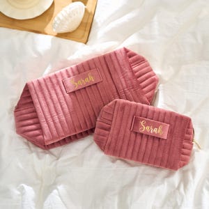May include: Two dusty rose velvet cosmetic bags with vertical quilted stitching. The larger bag has a rectangular label with the name "Sarah" in gold script. The smaller bag also has the name "Sarah" in gold script.