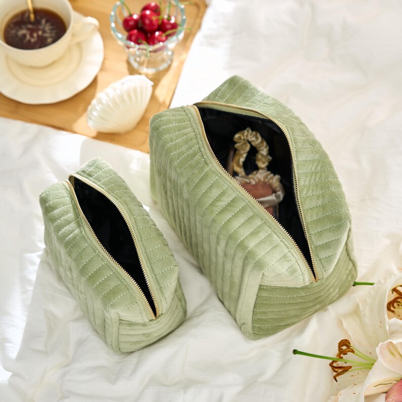 May include: Two sage green quilted velvet cosmetic bags with gold zippers. The bags are open, revealing a black interior. One bag is larger than the other. The bags are on a white surface.