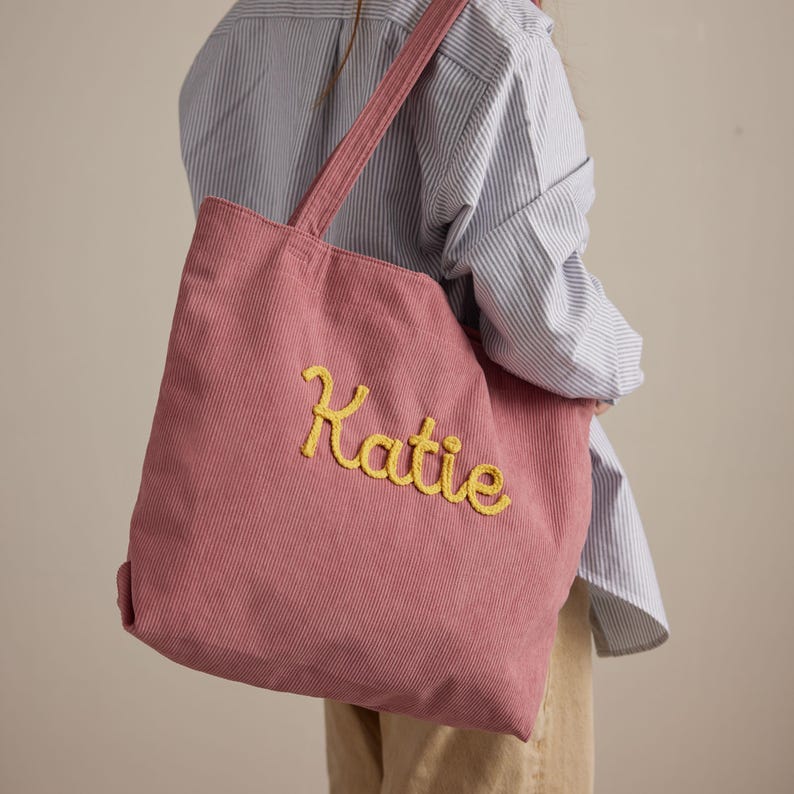 May include: A dusty rose corduroy tote bag with the name "Katie" embroidered in yellow script. The bag has matching straps and is being carried over the shoulder. The background includes a striped blue and white shirt.