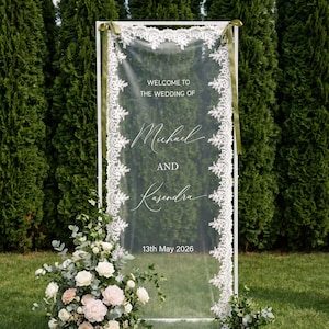 Könnte beinhalten: Transparente Hochzeitsanzeige mit weißer Spitzeneinfassung und grünen Bändern. Auf der Anzeige steht „Willkommen zur Hochzeit von Michael und Kasendra 13. Mai 2026“. Blumengestecke aus weißen und rosa Rosen befinden sich am Fuß.