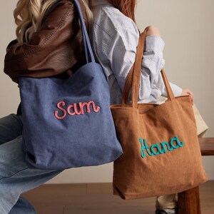 May include: Two corduroy tote bags, one blue and one brown, with embroidered names. The blue bag has "Sam" in pink, and the brown bag has "Hana" in teal. Both bags have long handles.