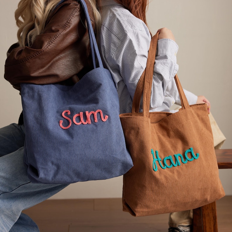 May include: Two corduroy tote bags, one blue and one brown, with embroidered names. The blue bag has "Sam" in pink, and the brown bag has "Hana" in teal. Both bags have long handles.