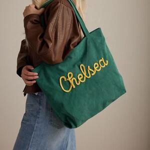 May include: A green corduroy tote bag with the name "Chelsea" embroidered in yellow script. The bag has matching green handles and is being held by a person wearing a brown leather jacket and blue jeans.