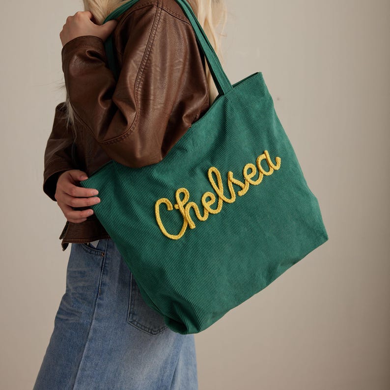 May include: A green corduroy tote bag with the name "Chelsea" embroidered in yellow script. The bag has matching green handles and is being held by a person wearing a brown leather jacket and blue jeans.
