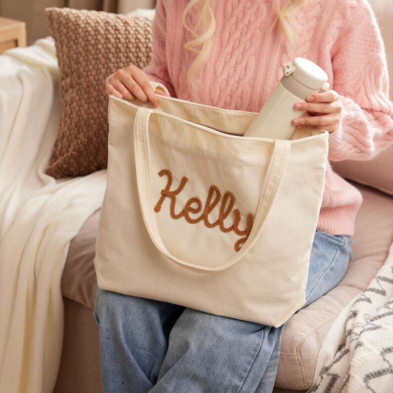 May include: Cream-colored corduroy tote bag with the name "Kelly" embroidered in brown script. The bag has a matching handle and is held by a person wearing a pink sweater. A white water bottle is inside the bag.