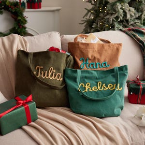 May include: Three corduroy tote bags in olive green, tan, and teal, each embroidered with a name in a contrasting color: Julia, Hana, and Chelsea. The bags are on a beige couch with wrapped gifts nearby, suggesting a holiday theme.