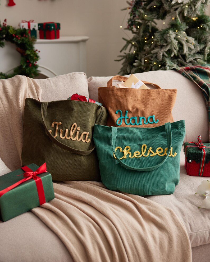 May include: Three corduroy tote bags in olive green, tan, and teal, each embroidered with a name in a contrasting color: Julia, Hana, and Chelsea. The bags are on a beige couch with wrapped gifts nearby, suggesting a holiday theme.