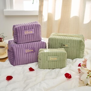 May include: Four corduroy cosmetic bags in shades of purple and green, with gold zippers. The bags are rectangular and have the names "Olivia" and "Laura" in gold lettering. The bags are arranged on a white surface.