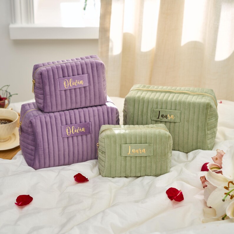 May include: Four corduroy cosmetic bags in shades of purple and green, with gold zippers. The bags are rectangular and have the names "Olivia" and "Laura" in gold lettering. The bags are arranged on a white surface.