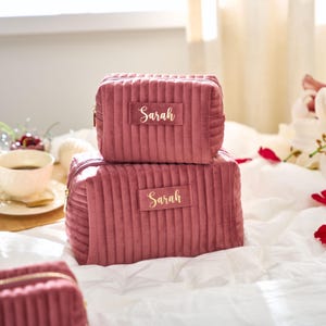 May include: Two stacked, dusty rose-colored, corduroy cosmetic bags with gold zippers. Each bag has a rectangular label with the name "Sarah" in gold lettering. The bags are on a white bedspread.