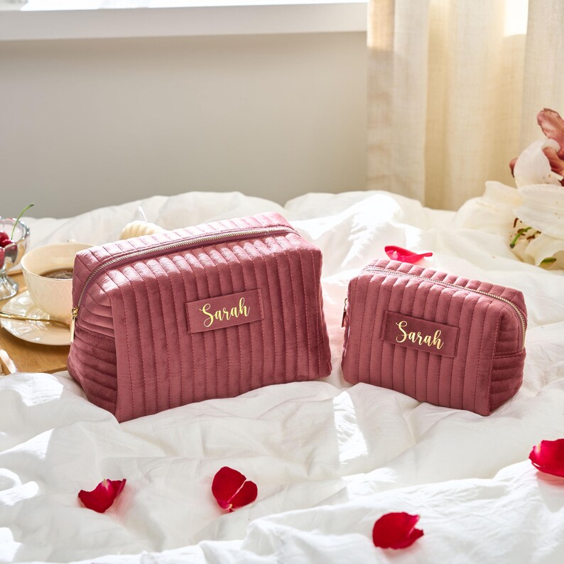 May include: Two dusty rose velvet cosmetic bags with gold zippers and the name "Sarah" in gold lettering. The bags have a ribbed design and are on a white bed with rose petals.