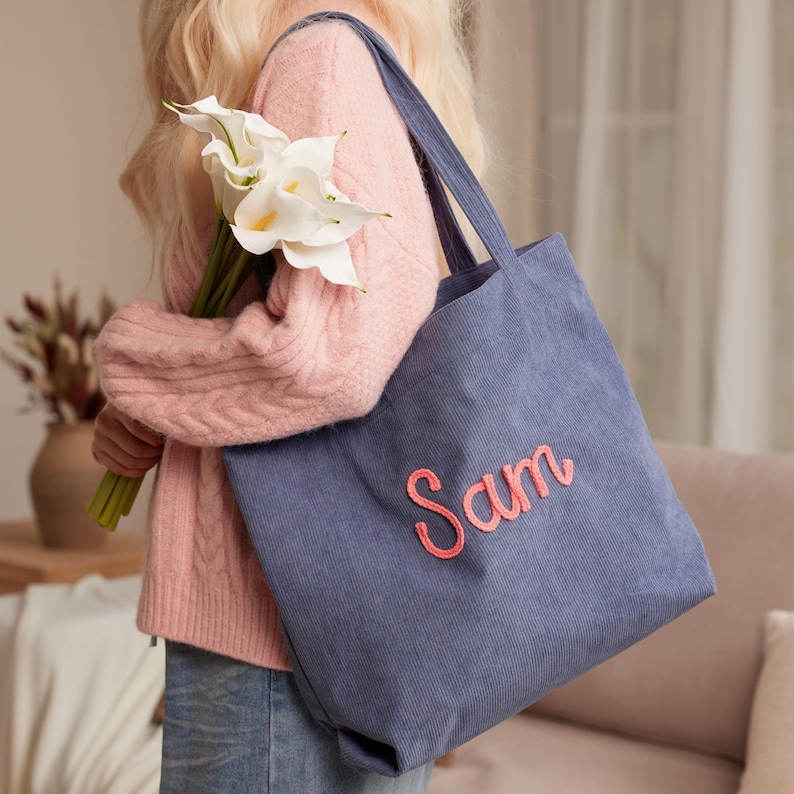 May include: A blue corduroy tote bag with the name "Sam" embroidered in pink. The bag has a rectangular shape and long straps. The person is holding a bouquet of white calla lilies.