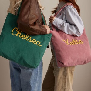 May include: Two corduroy tote bags, one emerald green and one dusty rose, are displayed. The green bag has "Chelsea" embroidered in yellow, while the pink bag has "Katie" embroidered in yellow. Both bags have a simple, rectangular design.