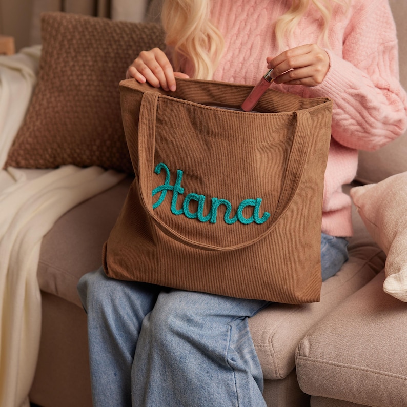 May include: A brown corduroy tote bag with the name "Hana" embroidered in teal script. The bag has long handles and is open, revealing a lipstick inside. The person is wearing a pink sweater and blue jeans.
