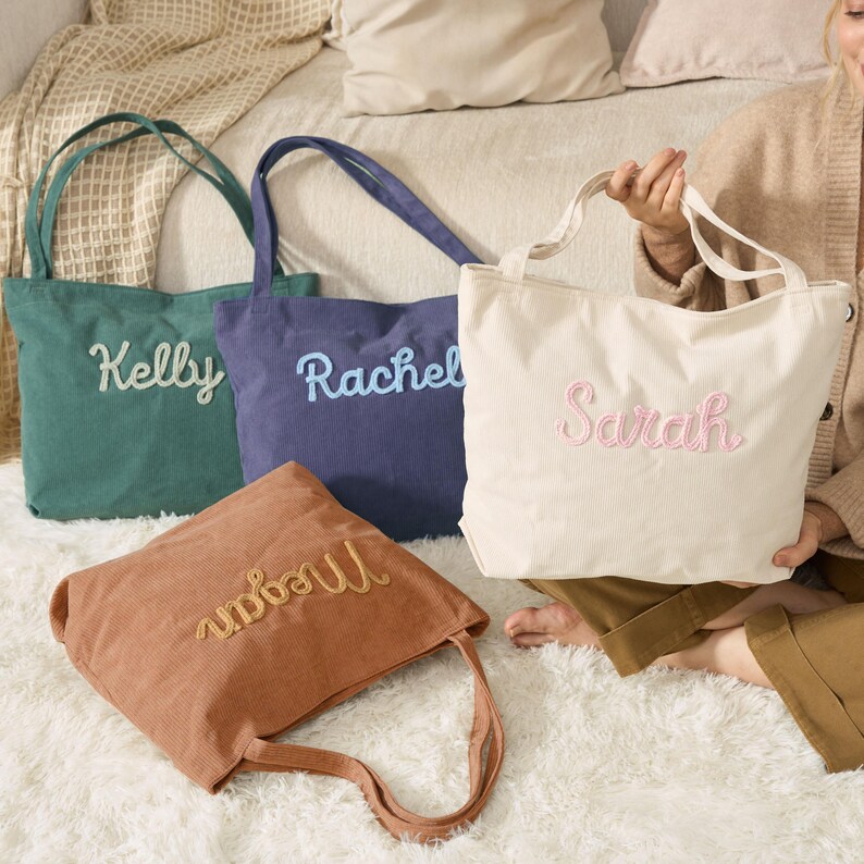 May include: Four corduroy tote bags in various colors: green, blue, tan, and cream. Each bag features an embroidered name: Kelly, Rachel, Megan, and Sarah. The bags have long handles and are displayed on a white fluffy surface.