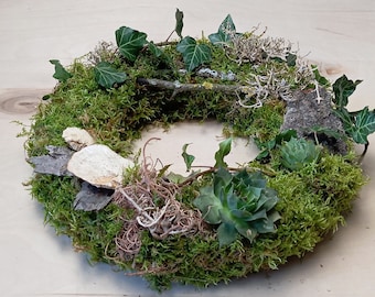 Op de afbeelding: Een ronde krans gemaakt van groen mos, voorzien van donkergroene klimopbladeren, kleine vetplantjes, stukjes boomschors, lichtgekleurde boomzwammen en dunne bruine takjes in een natuurlijke, textuurrijke compositie.