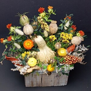 Könnte beinhalten: Eine rechteckige Holzkiste mit einem Trockenblumenarrangement, bestehend aus einem hellen Kürbis in der Mitte, orangefarbenen und gelben Strohblumen, getrockneten Mohnkapseln, grünen Eukalyptusblättern, weißen Hasenschwanzgräsern und einem braunen Kiefernzapfen.