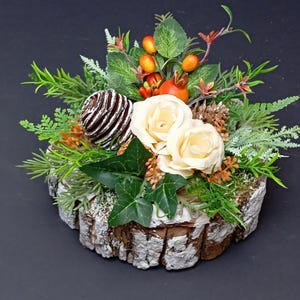 May include: A decorative arrangement featuring two cream-colored roses, a brown pinecone, green ivy leaves, and orange berries set atop a circular piece of natural wood bark. The floral display includes various green foliage and pine needles.