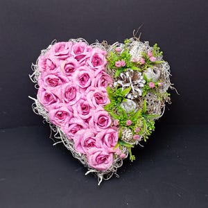 Pode incluir: Um arranjo decorativo em forma de coração com um grupo de rosas de tecido rosa no lado esquerdo, contrastado com folhagem verde, pequenos botões cor-de-rosa, pinhas prateadas e fibras naturais secas preenchendo o lado direito do coração.