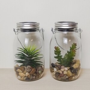 May include: Two clear glass jars with metal lids and wire handles. Each jar contains small rocks and a green artificial plant. The jars have solar powered lights on the lids.