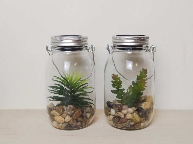 May include: Two clear glass jars with metal lids and wire handles. Each jar contains small rocks and a green artificial plant. The jars have solar powered lights on the lids.
