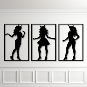May include: Three framed black silhouette art pieces of women with horns, each in a different pose. The art is displayed on a white wall above a wooden floor. The frames are black and rectangular.
