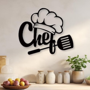 Puede incluir: Arte de pared de metal negro con la palabra "Chef" en una fuente estilizada, acompañada de un gorro de chef y una espátula. La obra de arte está montada en una pared de color claro, con una encimera de cocina y varios artículos de cocina en primer plano.