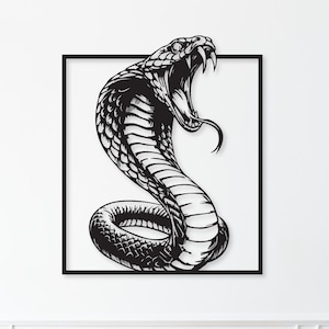 Op de afbeelding: Zwart-wit cobra slang muurkunst in een zwart frame. De slang wordt afgebeeld met open mond, zichtbare giftanden en uitgestoken tong. Het kunstwerk is gemonteerd op een witte muur boven een houten kast met twee lades en een deur.